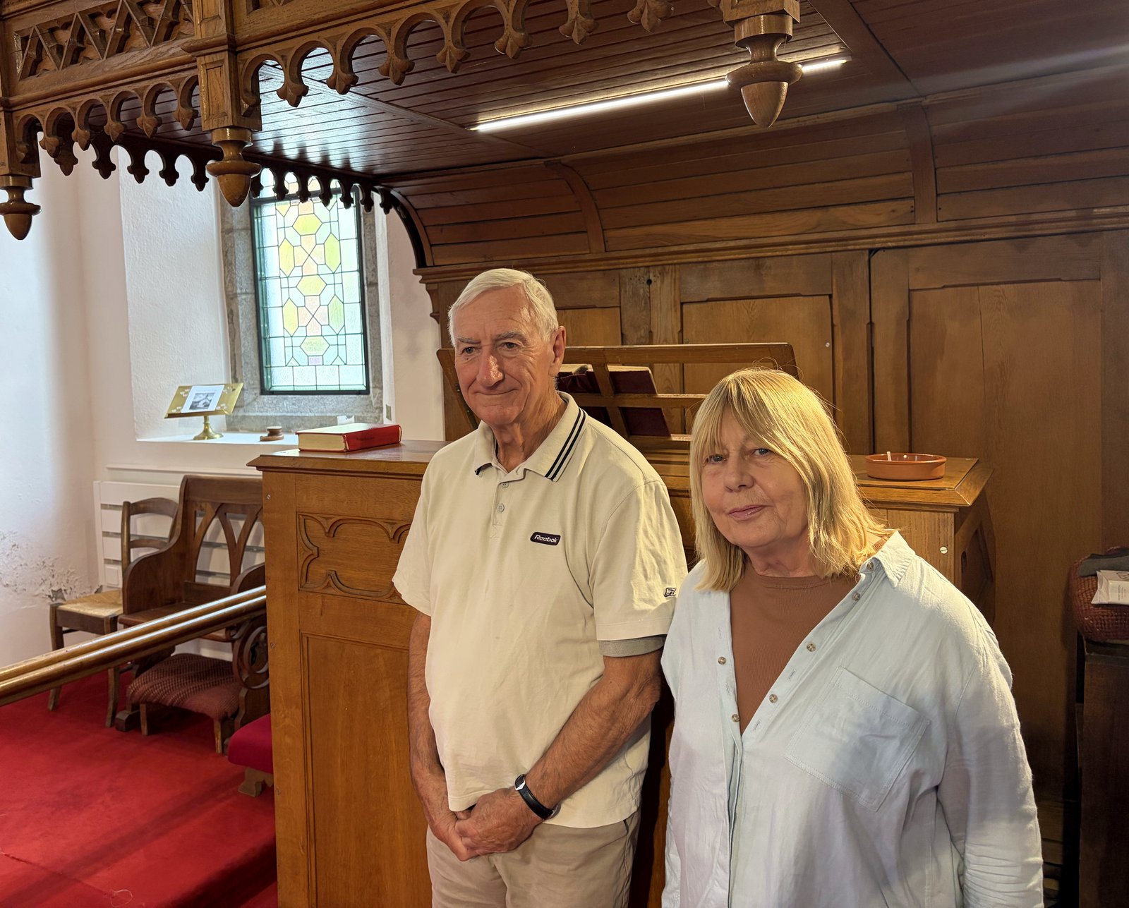 Peter and Carol Murray standing together inside St Peter's church, Château-d'Œx, beside the carved wooden pulpit