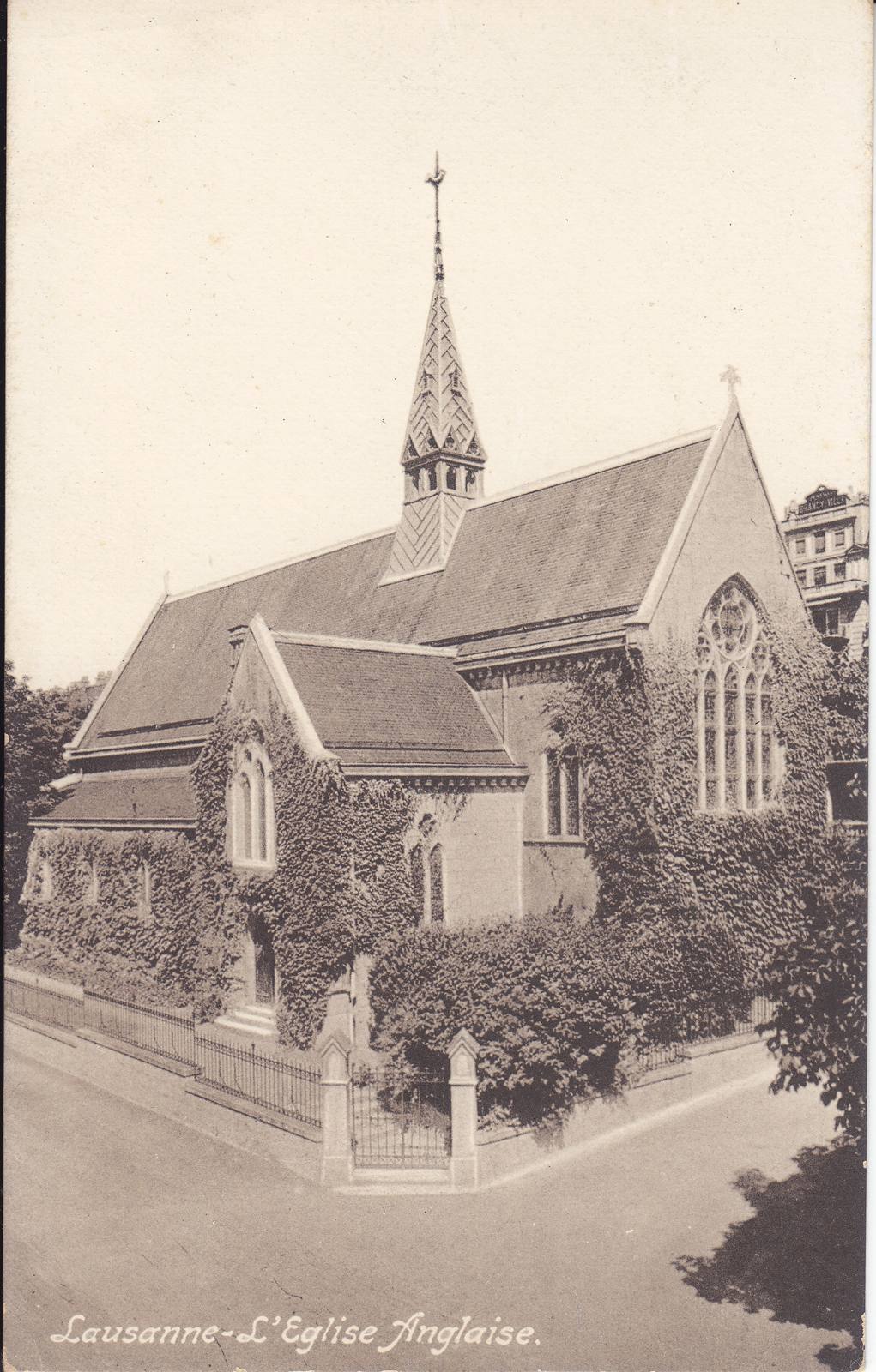 Christ Church, Lausanne, covered in ivy, seen from a street corner behind railings, with a tall spired bell turret