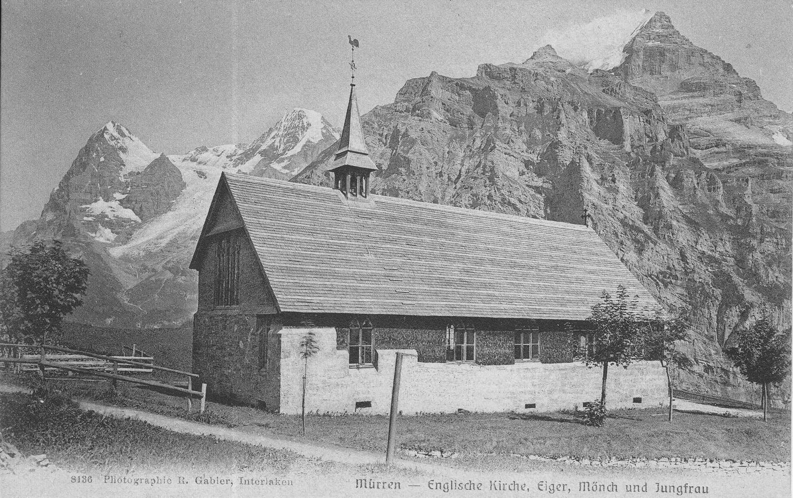The English church at Mürren, a long building with a steep roof and a small spire, with the Eiger, Mönch and Jungfrau behind