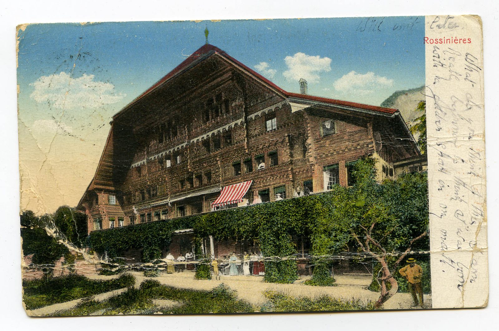 A hand-coloured postcard of the Grand Chalet at Rossinière, a vast carved wooden building with guests at tables in the garden, with a handwritten message down the right margin