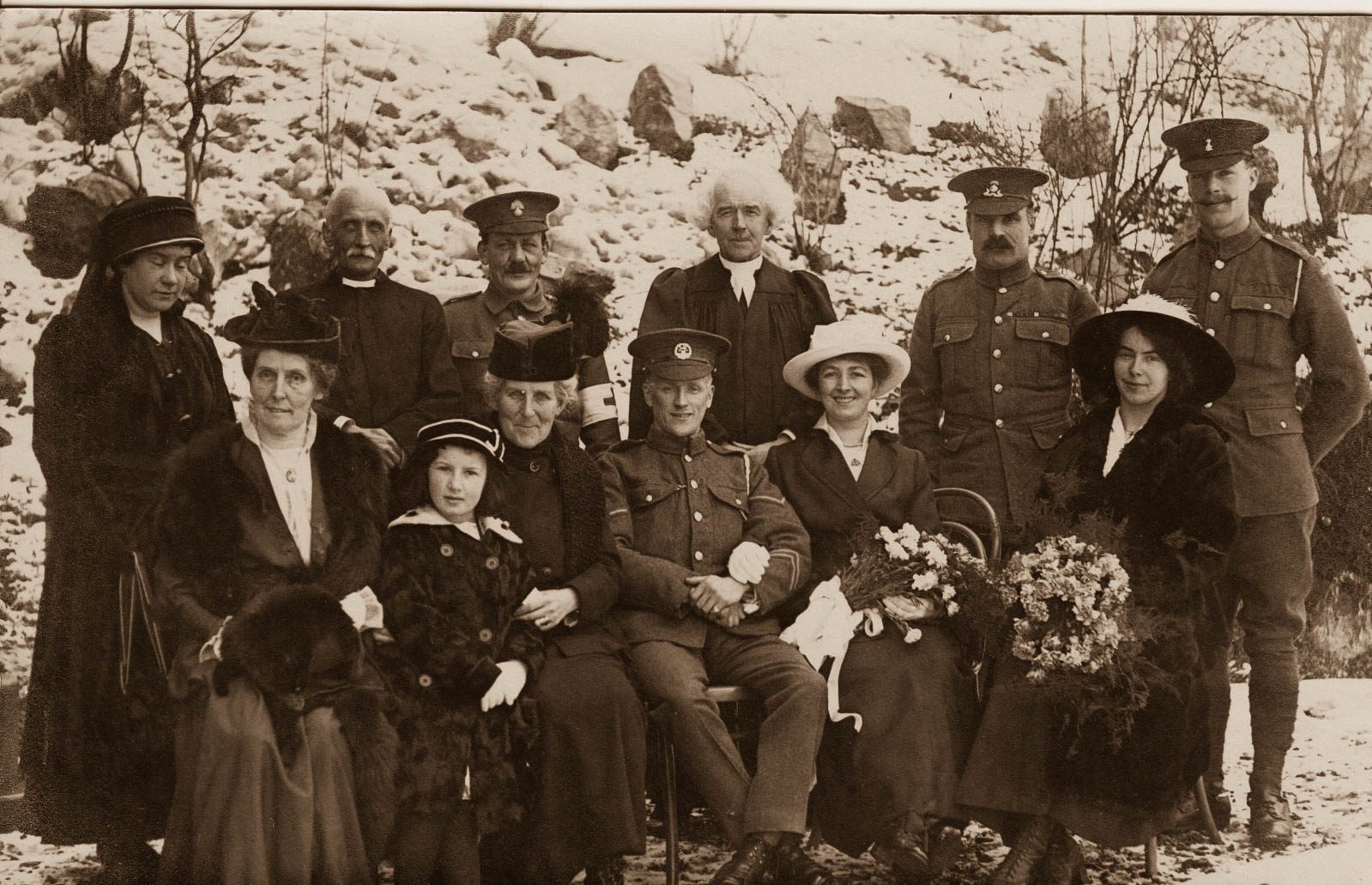 A wedding group photographed in the snow at Château-d'Œx: a seated soldier and his bride surrounded by women in furs and wide hats, soldiers standing behind, and two clergymen