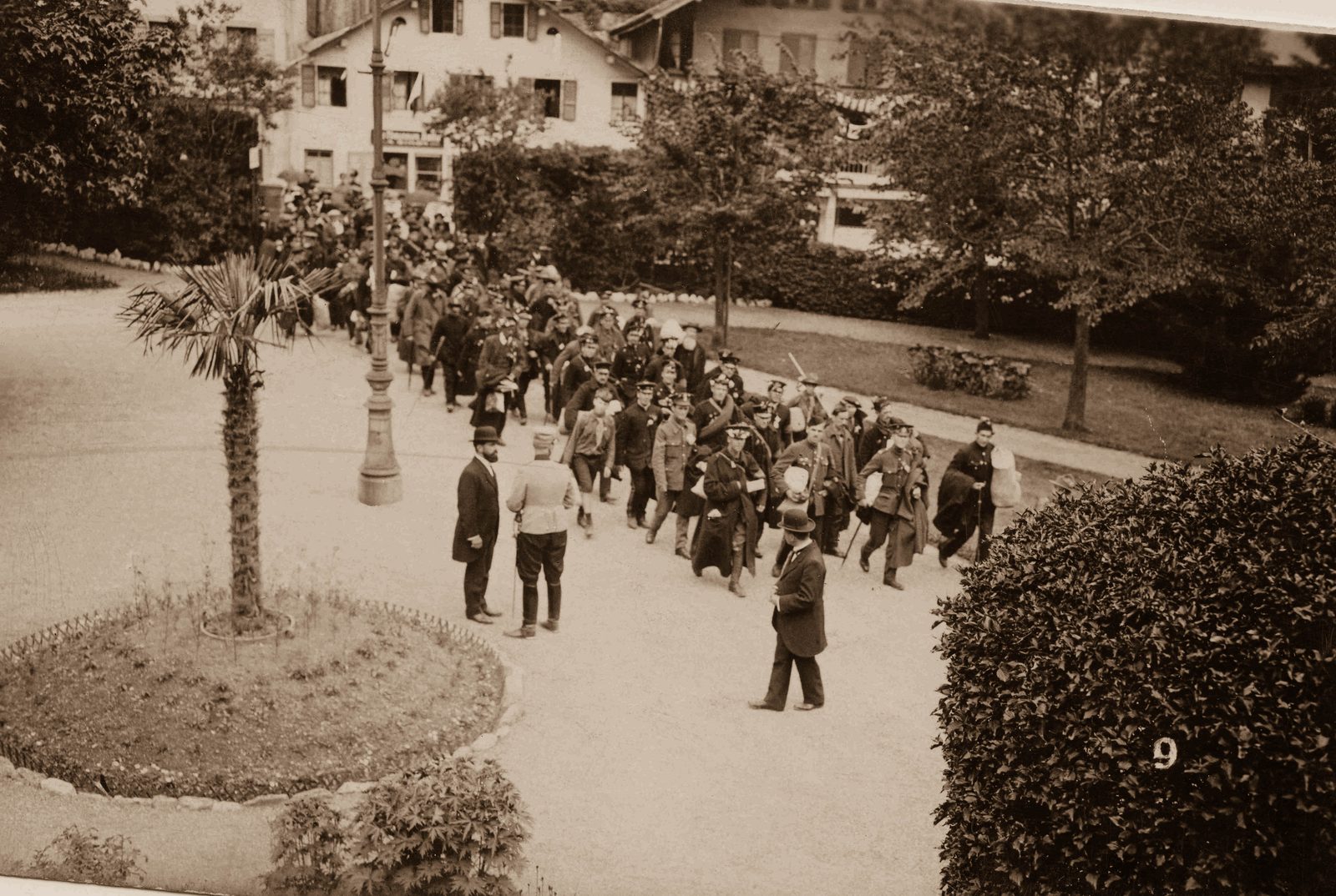 A long column of British internees in greatcoats and caps walks up a gravel path through hotel gardens at Château-d'Œx, watched by a crowd and by men in bowler hats