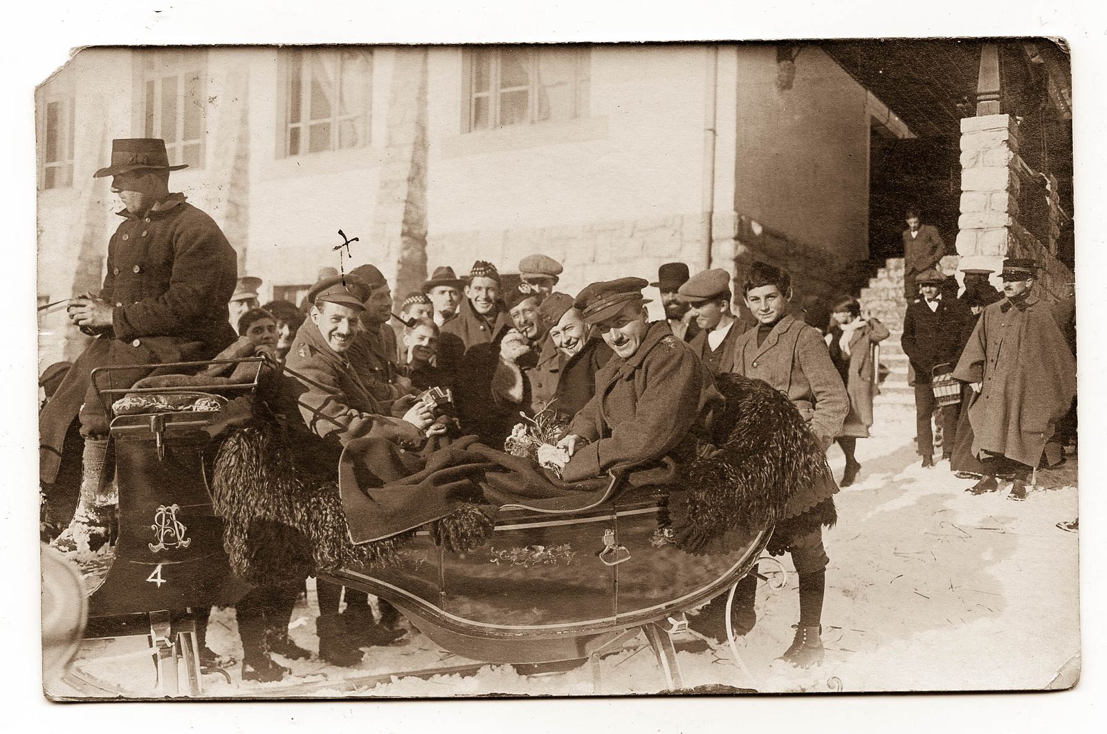 A group of grinning British internees packed into a horse sledge with fur-trimmed sides in the snow at Château-d'Œx, watched by village boys