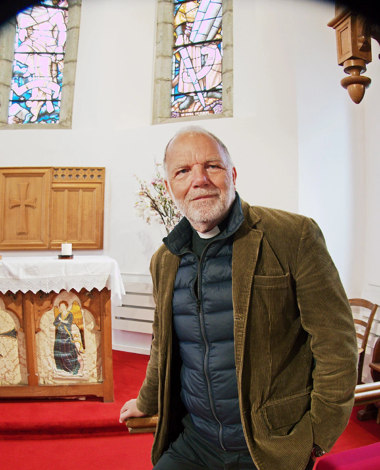 Revd Paul Cowley MBE standing in St. Peter's, Château-d'Oex, beside the altar with its painted angel panels, beneath the stained glass
