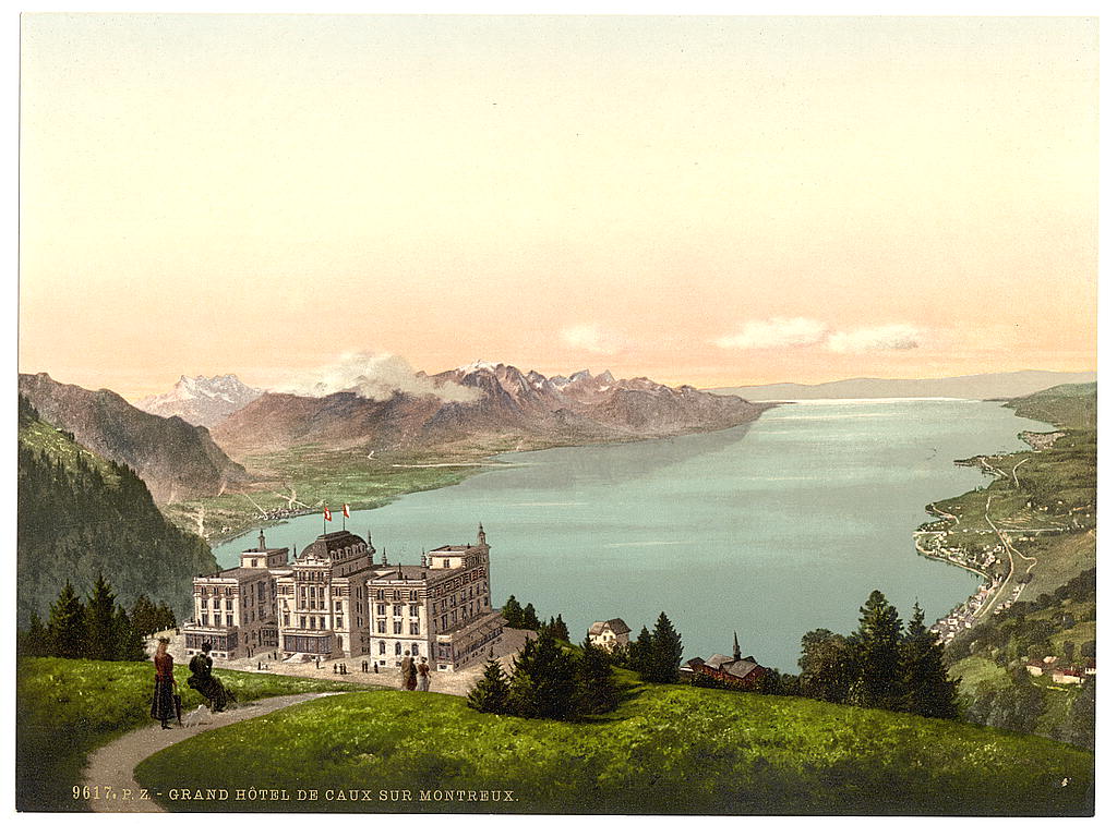 A large hotel on a green mountain terrace high above Lake Geneva, with two women in long dresses and a dog on a path in the foreground