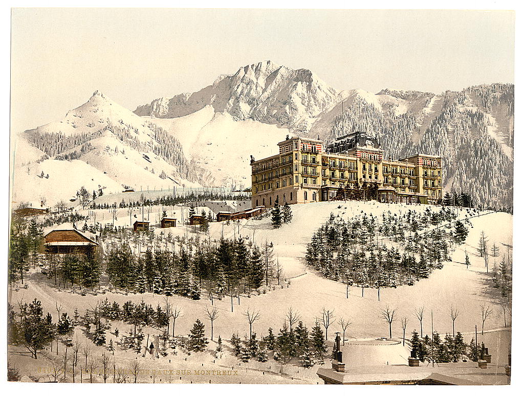 A large hotel standing in deep snow on a mountain terrace, with snow-covered peaks and forest behind it